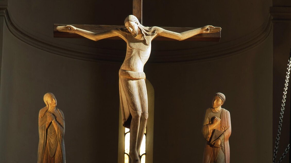 Ein Holzkreuz mit einer Skulptur von Jesus Christus, der gekreuzigt ist, steht im Mittelpunkt. Zwei Figuren stehen seitlich des Kreuzes in der Kirche. | © Abtei Seckau