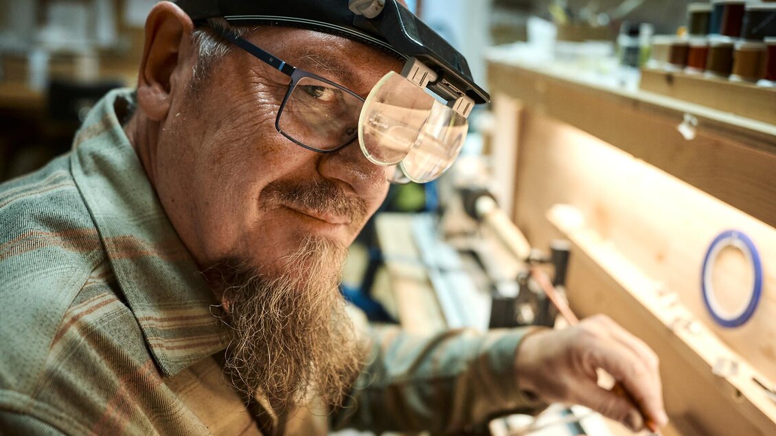 Ein älterer Mann mit einer Lupe arbeitet an einem handwerklichen Projekt. Im Hintergrund sind Werkzeuge und Materialien sichtbar.