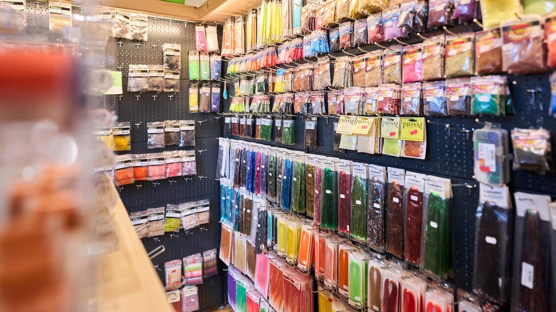 Ein gut sortierter Bastelshop mit einer Wand voller bunter Materialien in Verpackungen. Die Farben reichen von hellen bis dunklen Tönen und laden zum kreativen Arbeiten ein.