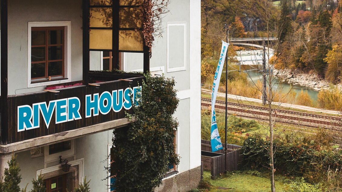 Ein Haus mit der Beschriftung "RIVER HOUSE" und Blick auf einen Fluss. Im Hintergrund sind Bäume und Gleise zu sehen, was eine ruhige Umgebung schafft.