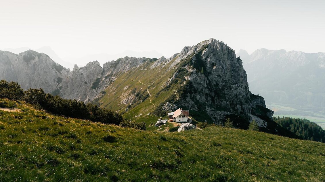 Eine beeindruckende Berglandschaft mit einer steilen Felsformation. Im Vordergrund steht ein kleines Haus inmitten von grünen Wiesen.