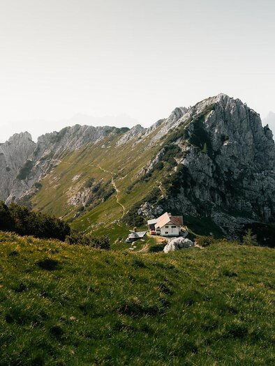 Eine beeindruckende Berglandschaft mit einer steilen Felsformation. Im Vordergrund steht ein kleines Haus inmitten von grünen Wiesen.