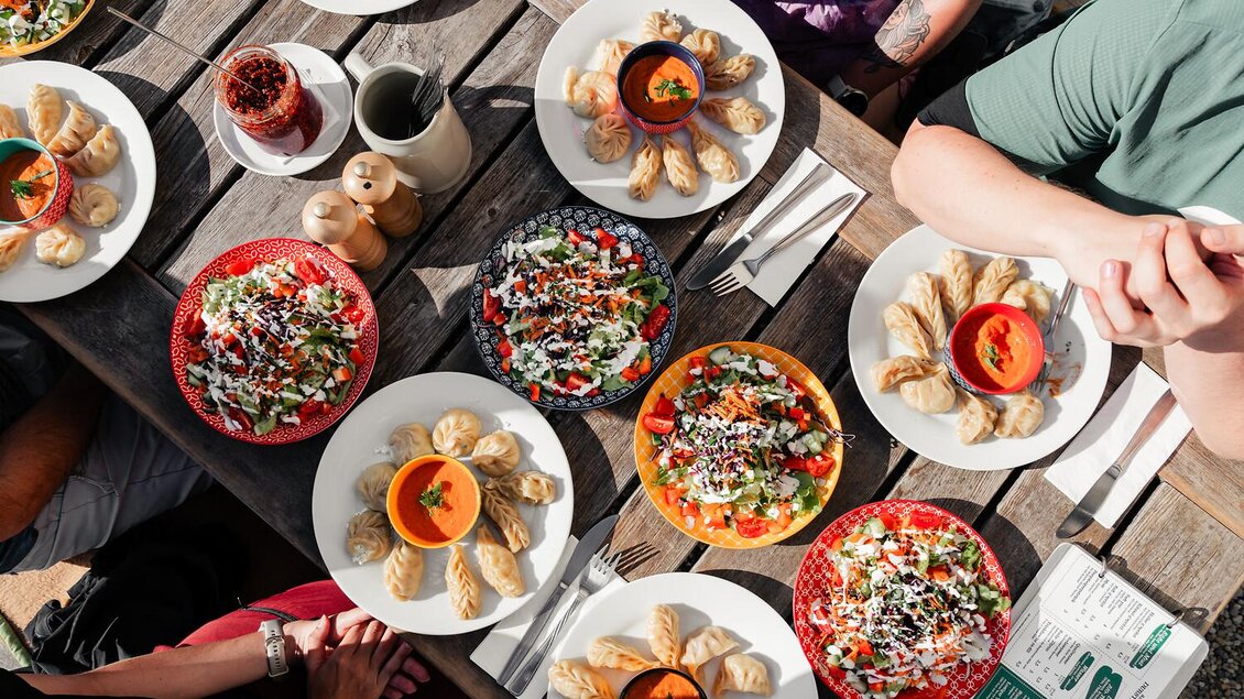 Ein Tisch voller farbenfroher Gerichte, darunter Salate und gefüllte Teigtaschen. Menschen sitzen zusammen und genießen das Essen in einer freundlichen Atmosphäre.