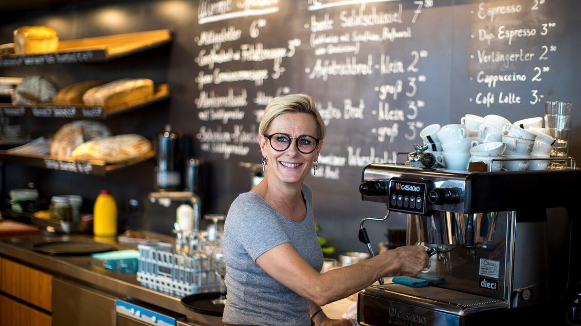 Eine lächelnde Barista steht an einer Espressomaschine in einem Café. Im Hintergrund sind Brote und eine Tafel mit Getränkepreisen zu sehen. | © Region Graz - Mias Photoart