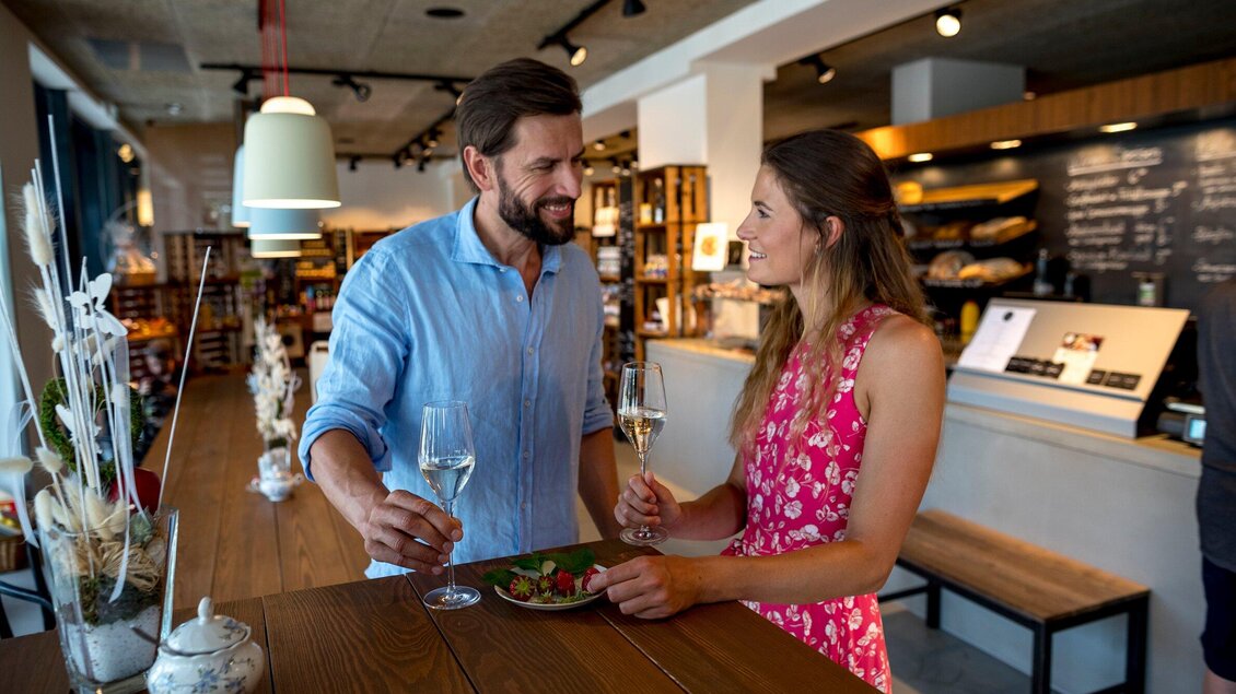 Ein Paar steht in einem modernen Café und prostet sich mit Gläsern Sekt zu. Auf dem Tisch liegt ein Teller mit leckerem Essen. | © Region Graz - Tom Lamm