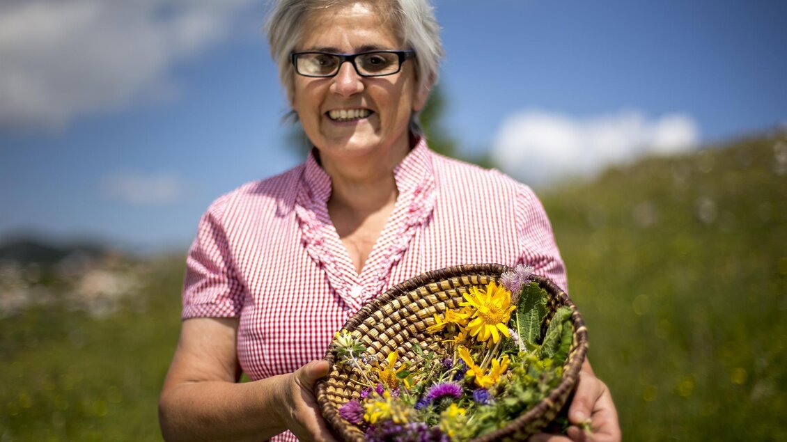 Eine lächelnde Frau hält einen Korb mit bunten Wildblumen in den Händen. Im Hintergrund sind grüne Wiesen und ein blauer Himmel zu sehen. | © Tom Lamm