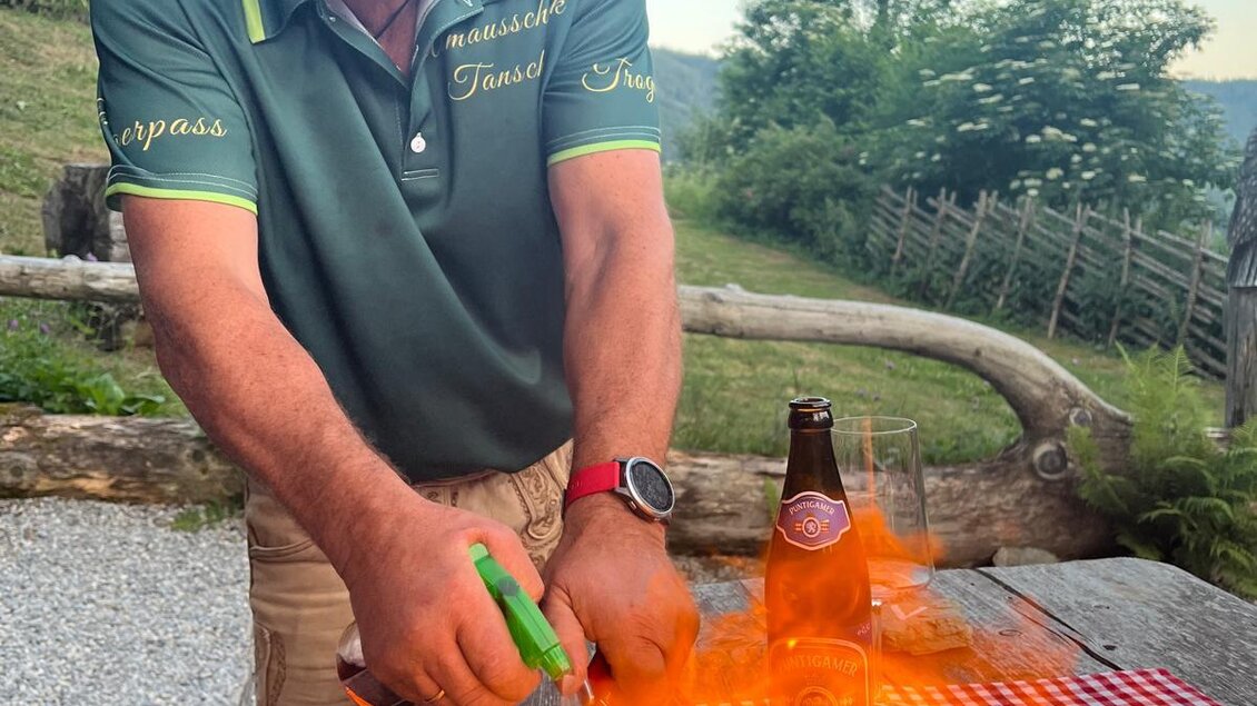 Ein Mann bereitet ein Gericht vor und zündet es an. Im Hintergrund sind eine schöne Landschaft und ein Bier sichtbar. | © Erwin Troger