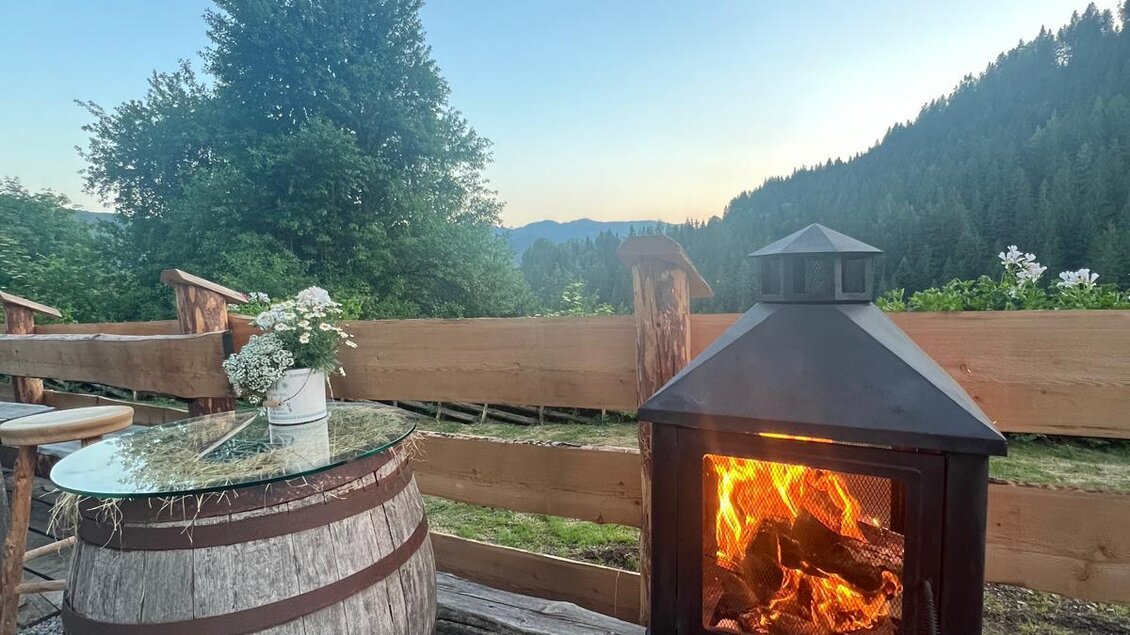 Eine gemütliche Terrasse mit einem Feuerofen und einer Holztonne. Im Hintergrund sind Bäume und Berge zu sehen, während der Himmel blau ist. | © Erwin Troger