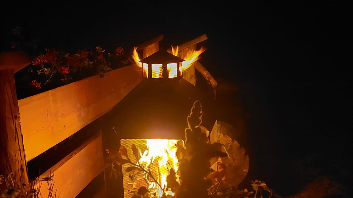 Eine gemütliche Feuerstelle leuchtet in der Dunkelheit. Umgeben von Pflanzen und einem Holzzaun schafft sie eine einladende Atmosphäre. | © Erwin Troger