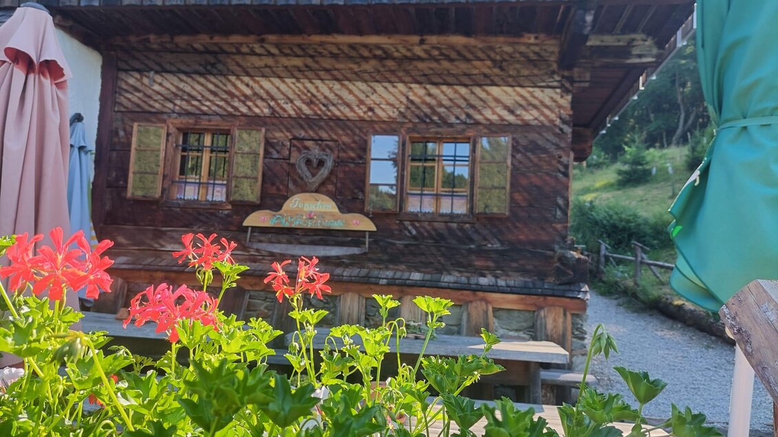 Eine gemütliche Hütte aus Holz mit bunten Fenstern und einem herzlichen Detail über der Tür. Im Vordergrund blühen rote und grüne Pflanzen. | © Erwin Troger