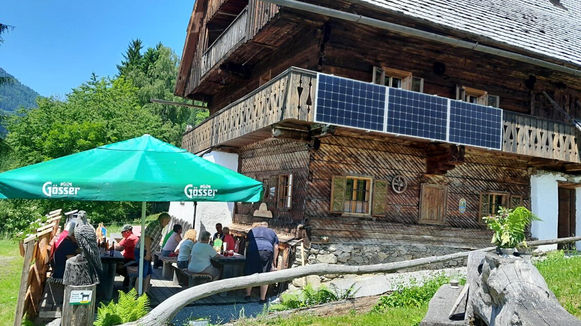 Ein traditionelles Holzhaus in der Natur mit einem grünen Sonnenschirm. Gruppen von Menschen sitzen draußen und genießen die Umgebung. | © Erwin Troger