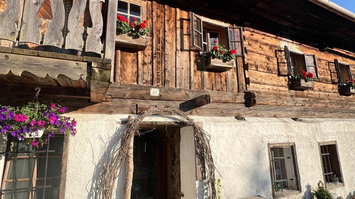 Ein traditionelles Holzhaus mit einem begrünten Eingangsbereich und Blumenkästen an den Fenstern. Die Fassade zeigt rustikale Details und klaren blauen Himmel im Hintergrund. | © Erwin Troger