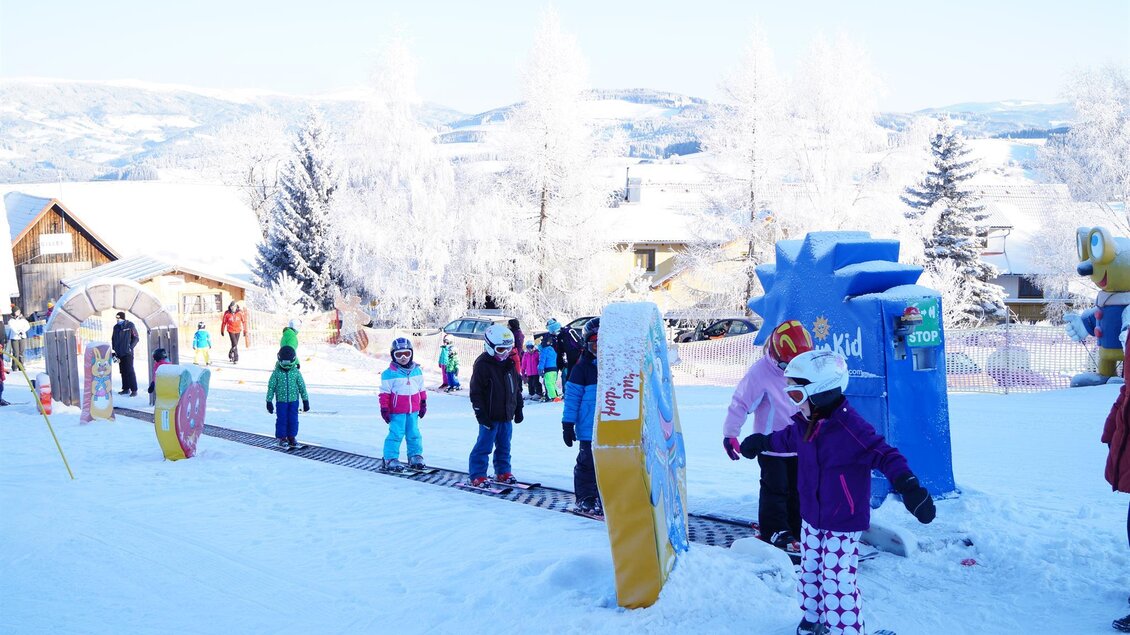 Eine verschneite Winterlandschaft mit vielen Kindern, die Ski fahren lernen. Im Hintergrund sind Häuser und Bäume zu sehen. | © Gasthof Almblick