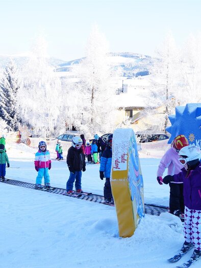 A snowy winter landscape with many children learning to ski. Houses and trees are visible in the background. | © Gasthof Almblick