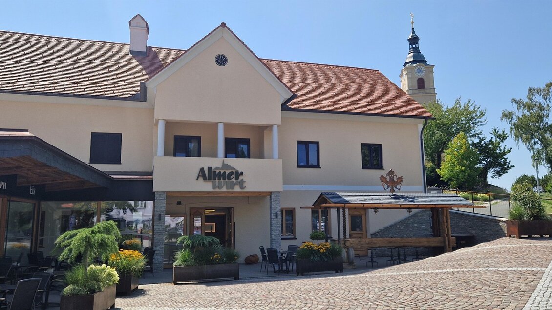 Ein hübsches Gebäude mit einem Restaurant und einer Terrasse im Freien. Im Hintergrund ist ein Kirchturm zu sehen, umgeben von Bäumen. | © Almer