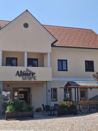 Ein hübsches Gebäude mit einem Restaurant und einer Terrasse im Freien. Im Hintergrund ist ein Kirchturm zu sehen, umgeben von Bäumen. | © Almer