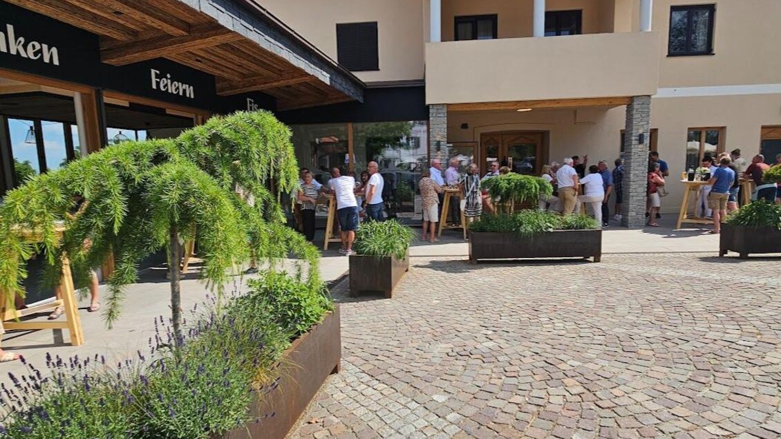 Ein geschäftiger Eingang zu einem Gebäude mit einem schönen, gepflasterten Platz. Im Vordergrund stehen dekorative Pflanzen und es sind viele Menschen zu sehen. | © Sophie Dekkers