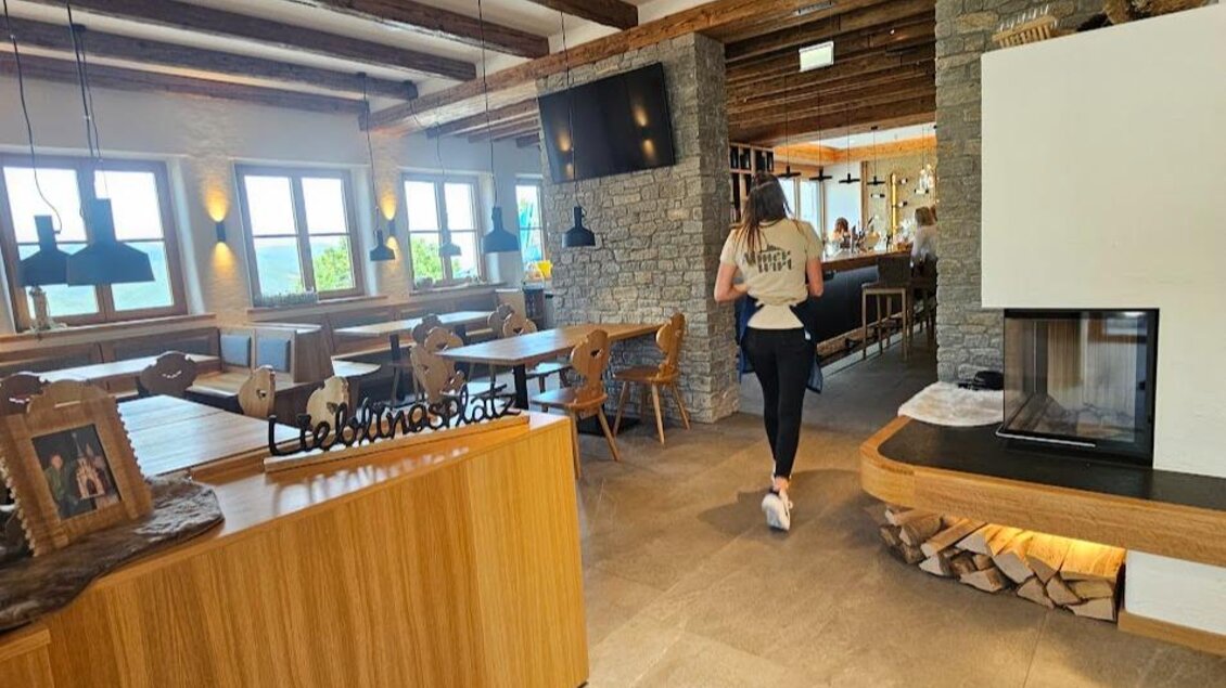 Ein modernes Restaurant mit Holzdecken und großen Fenstern. Eine Person geht durch den Raum, neben einem offenen Kamin und gemütlichen Tischen. | © Sophie Dekkers