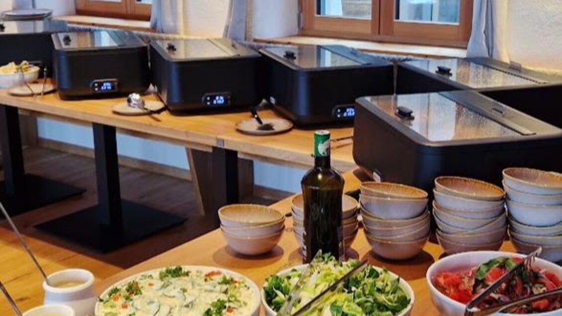 Ein Buffettisch mit verschiedenen Salaten und Beilagen. Im Hintergrund sind mehrere Heizplatten und große Fenster zu sehen. | © Sophie Dekkers