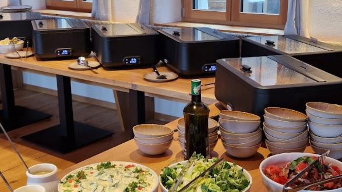 Ein gut gedeckter Tisch mit verschiedenen Salaten und Beilagen. Im Hintergrund sind große Fenster und eine gemütliche Atmosphäre zu sehen. | © Almerwirt