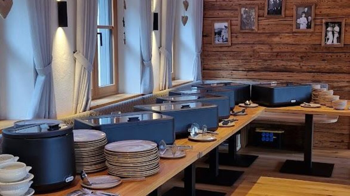 Ein gemütlicher Raum mit großen Holztischen und modernem Licht. Es gibt Geschirr und Buffetstationen für eine Mahlzeit. | © Almerwirt