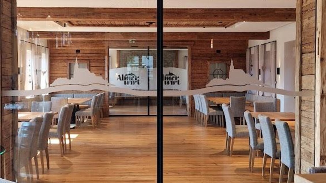 Ein modernes Restaurant mit Holzelementen und einem einladenden Innenraum. Die großen Glasschieben bieten einen Blick auf die Tische und Stühle im Raum. | © Almerwirt