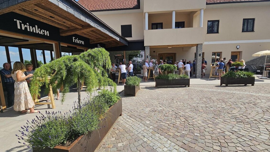 Eine charmante Weinstube mit einer langen Warteschlange von Gästen vor dem Eingang. Im Vordergrund sind blühende Pflanzen in modernen Blumentöpfen zu sehen. | © Almerwirt