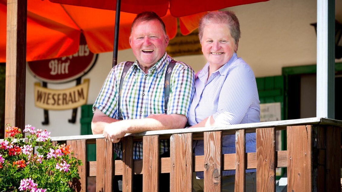 Zwei Personen lächeln auf der blumengeschmückten Terrasse einer Almhütte unter roten Sonnenschirmen | © TV Region Graz - René Vidalli