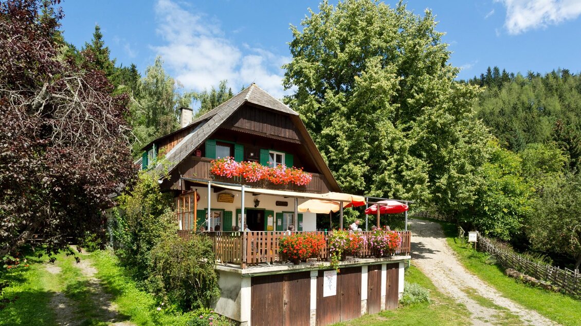 Traditionelles Holzhaus mit Blumenbalkon und Sonnenschirmen umgeben von Bäumen und Wiesen | © TV Region Graz - René Vidalli