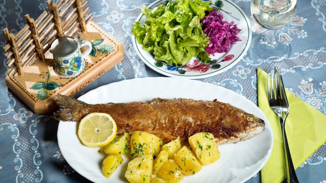 Ein gebratener Fisch liegt auf einem weißen Teller, umgeben von frischen Kartoffeln und einer Zitronenscheibe. Neben dem Teller befindet sich ein Salat mit grünem Blattgemüse und Rotkohl. | © TV Region Graz - René Vidalli