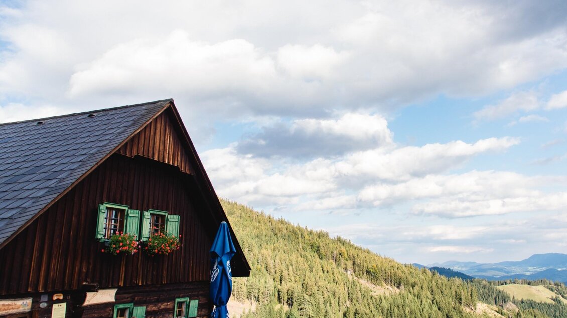 Ein malerisches Berghaus mit einem schrägen Dach und grünen Fensterläden. Im Hintergrund erstrecken sich die bewaldeten Hügel und der Himmel zeigt sanfte Lichtstrahlen. | © TV Region Graz - Mias Photoart