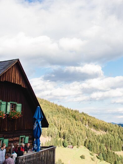 Ein malerisches Berghaus mit einem schrägen Dach und grünen Fensterläden. Im Hintergrund erstrecken sich die bewaldeten Hügel und der Himmel zeigt sanfte Lichtstrahlen. | © TV Region Graz - Mias Photoart