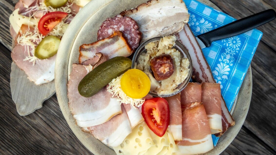 Ein rustikeller Teller mit verschiedenen Fleischsorten, Gurken und Tomaten. Im Hintergrund sind belegte Brötchen auf einem Holzbrett zu sehen. | © TV Region Graz -  Lunghammer