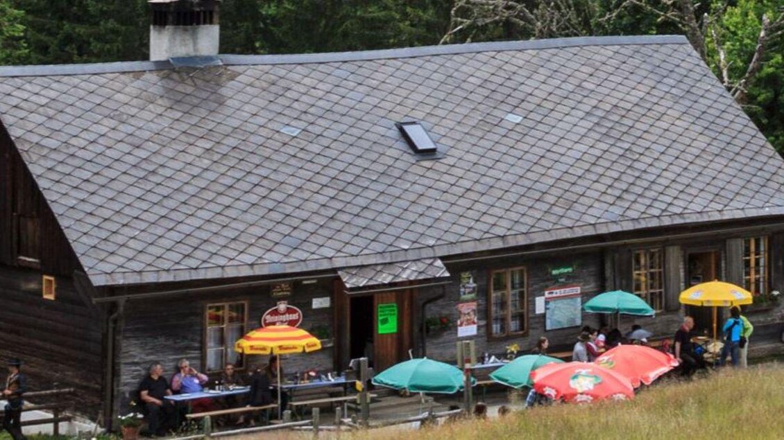 Eine gemütliche Holzhütte in der Natur mit Gästen, die draußen sitzen. Umbrella Tische laden zum Verweilen ein. | © Almhütte Wirtbartl