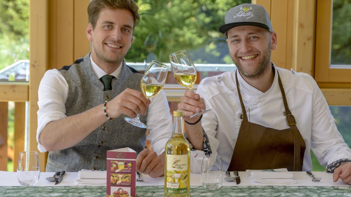 Zwei Männer stoßen mit Gläsern an, während sie an einem gedeckten Tisch sitzen. Im Hintergrund sind grüne Bäume und Tageslicht sichtbar. | © Almidylle Sabathy
