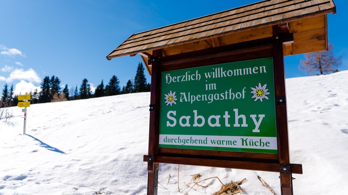 Ein Willkommensschild zum Alpengasthof Sabathy in verschneiter Landschaft. Darauf steht "Herzlich willkommen im Alpengasthof Sabathy" und "durchgehend warme Küche". | © Almidylle Sabathy