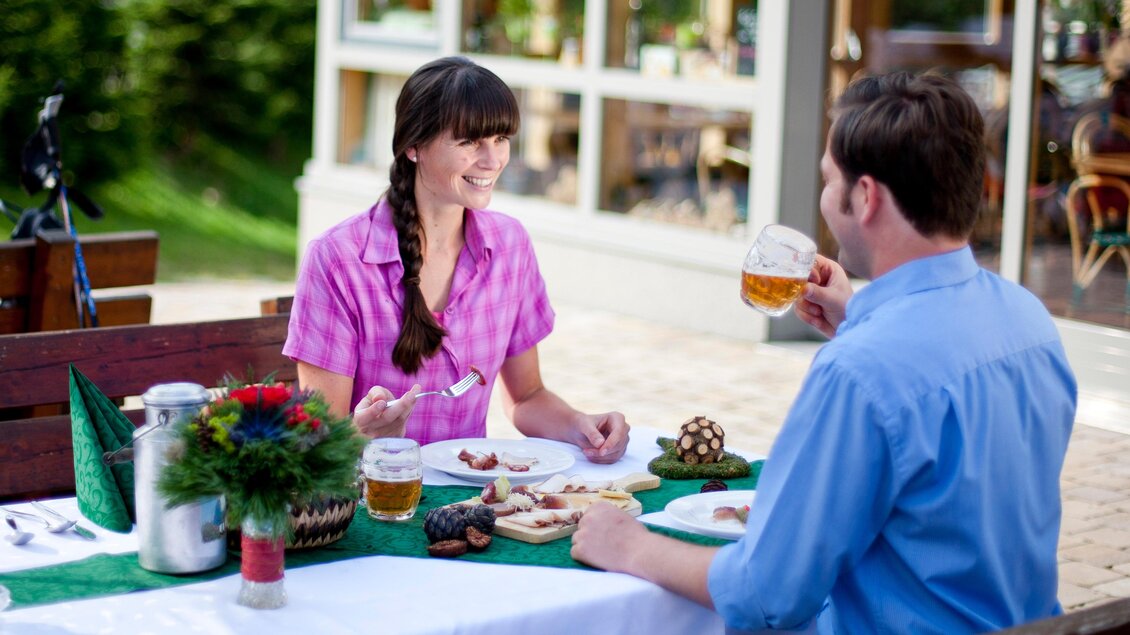 Ein Paar sitzt an einem Tisch im Freien und genießt eine Mahlzeit zusammen. Sie lachen und haben Getränke vor sich. | © Almidylle Sabathy
