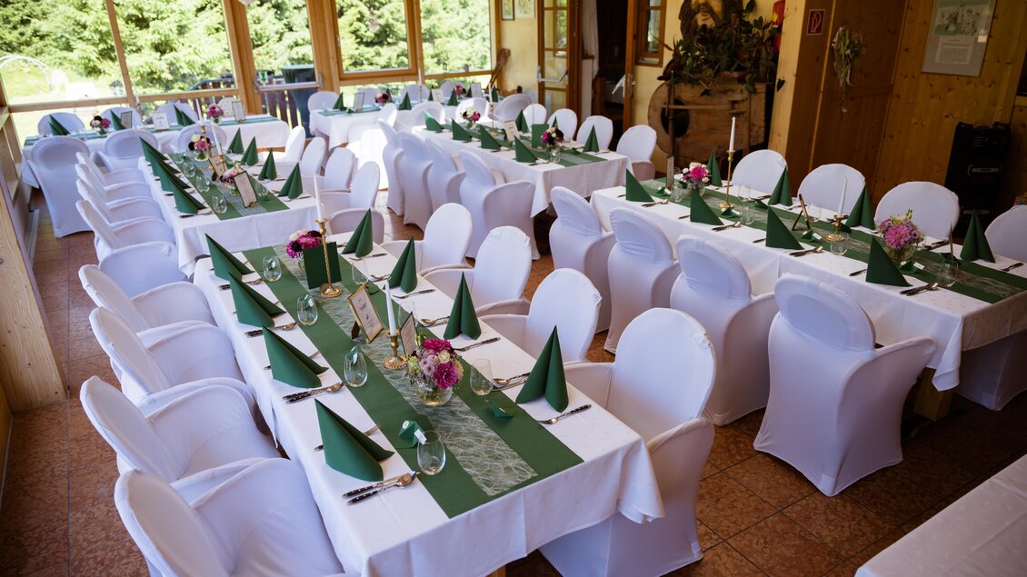 Ein festlich gedeckter Raum mit weißen Tischen und Stühlen. Jedes Tisch ist mit grünen Servietten und Blumen dekoriert. | © Almidylle Sabathy