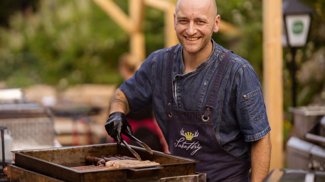 Ein lächelnder Koch grillt Fleisch auf einem Grill. Er trägt eine blaue Schürze und wirkt freundlich und einladend. | © Almidylle Sabathy
