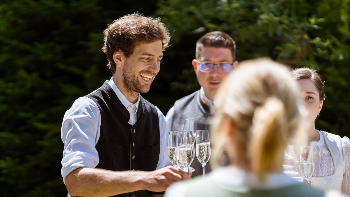 Eine fröhliche Außenaufnahme einer Feier mit Freunden. Ein Mann in traditioneller Kleidung hebt ein Glas und lächelt. | © Almidylle Sabathy
