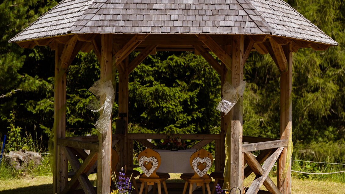 Ein romantisches Gartenhaus aus Holz, umgeben von Bäumen. Zwei Stühle mit Herzmotiven sind in der Mitte platziert, bereit für eine Feier. | © Almidylle Sabathy
