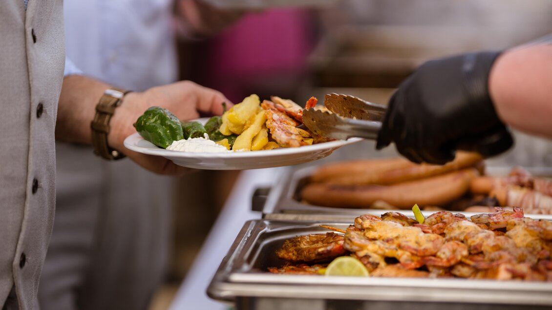 Ein Teller mit Gegrilltem, Garnelen und Beilagen wird serviert. Im Hintergrund sind weitere Speisen und eine Person zu sehen. | © Almidylle Sabathy