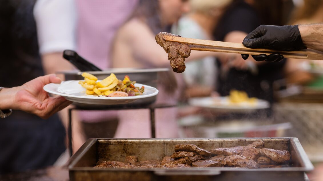 Ein Grillfest mit leckerem Fleisch und Beilagen wird serviert. Eine Hand reicht einen Teller mit Pommes und Fleisch. | © Almidylle Sabathy