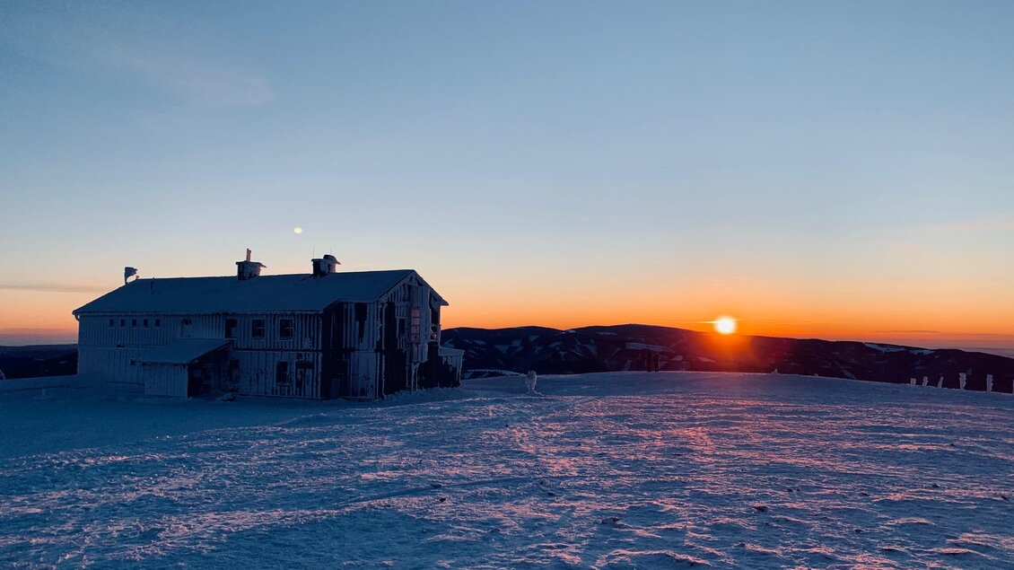 Ein Schneelandschaft mit einem weißen Haus im Vordergrund. Die Sonne geht am Horizont auf und färbt den Himmel in warmen Tönen. | © Gemeinde Rettenegg