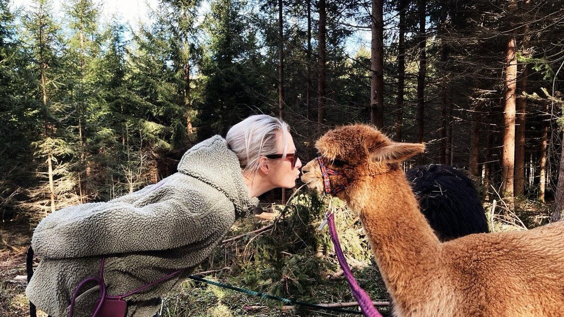 Eine Frau küsst ein Alpaka in einem Wald. Im Hintergrund sind hohe Bäume zu sehen. | © Sandrina Attems-Wimpffen