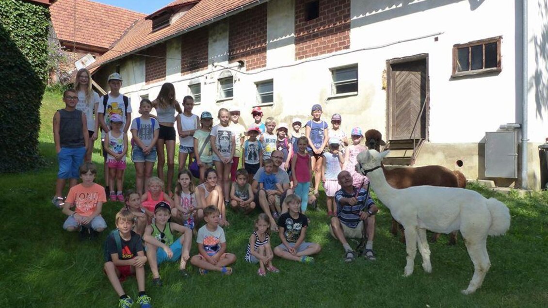 Eine Gruppe von Kindern steht auf einer Wiese vor einem Bauernhaus. Einige Tiere sind ebenfalls im Bild, darunter ein Lama und ein Pony. | © Alpakas Bratschitz