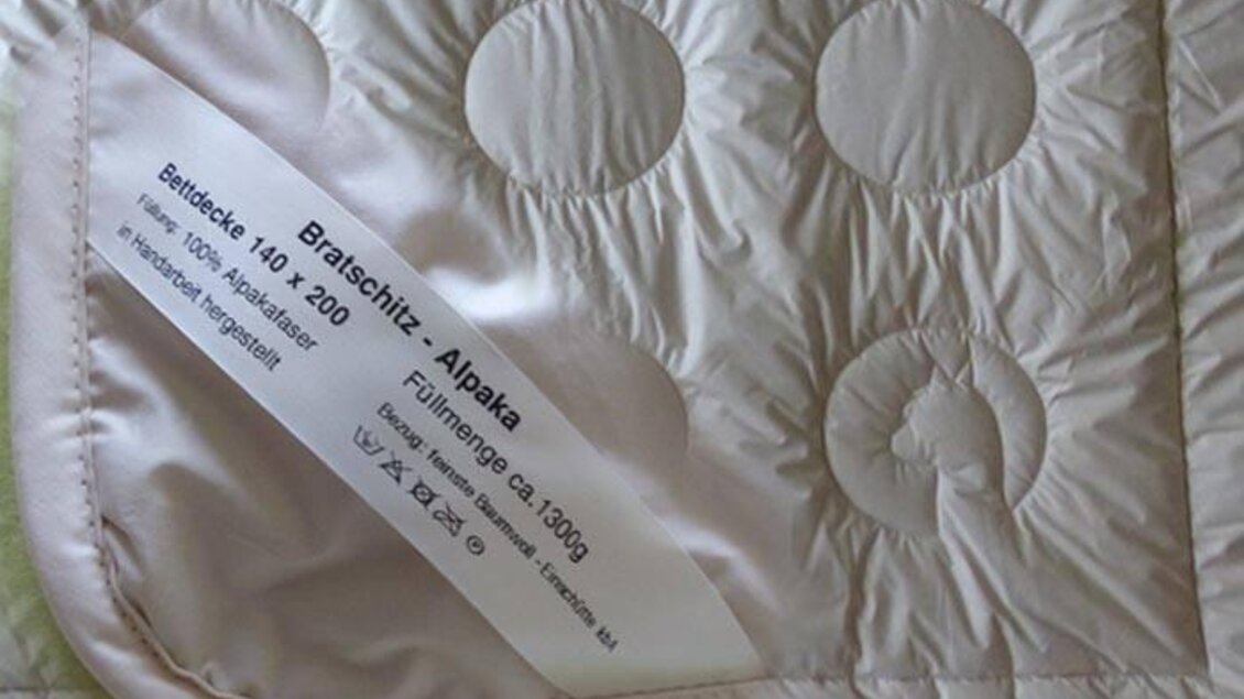 Eine weiche, hellrosa Bettdecke mit runden Mustern. Auf dem Etikett sind Informationen über das Material und die Maße angegeben. | © Alpakas Bratschitz