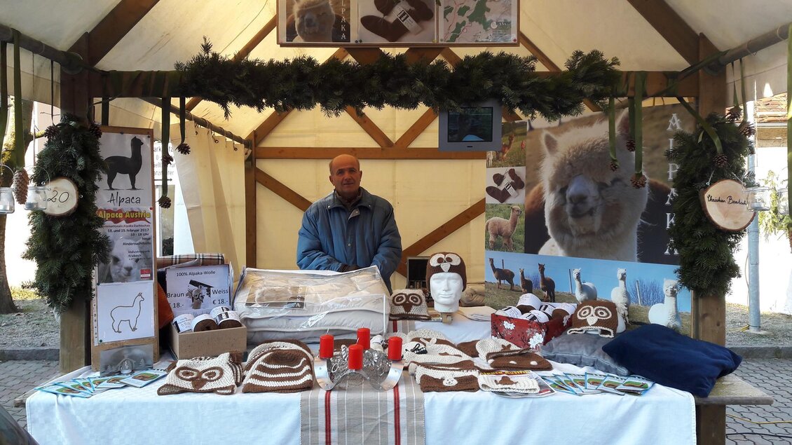 Ein Verkaufsstand mit handgefertigten Produkten aus Wolle und einem Verkäufer. Im Hintergrund sind Bilder von Tieren und dekorative Elemente zu sehen. | © Alpakas Bratschitz