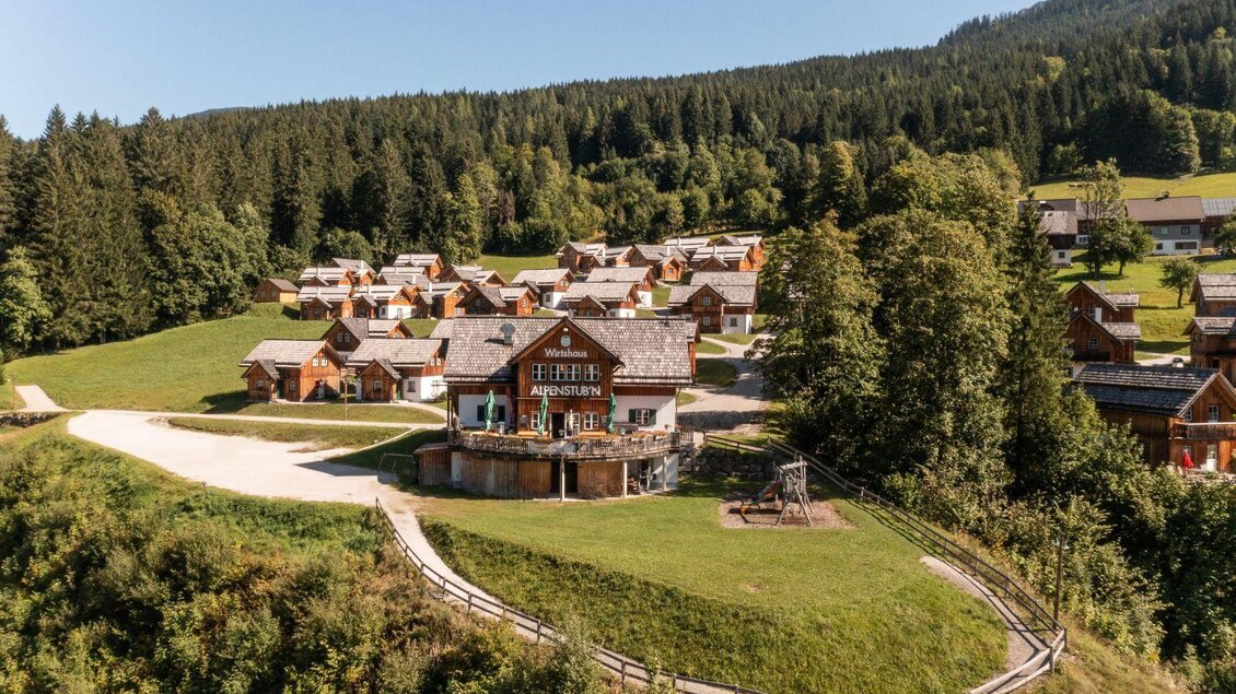 Eine malerische Landschaft mit Holzchalets inmitten von Bäumen. Im Vordergrund befindet sich ein Spielplatz und ein offener Bereich. | © Gerhard Wolkersdorfer