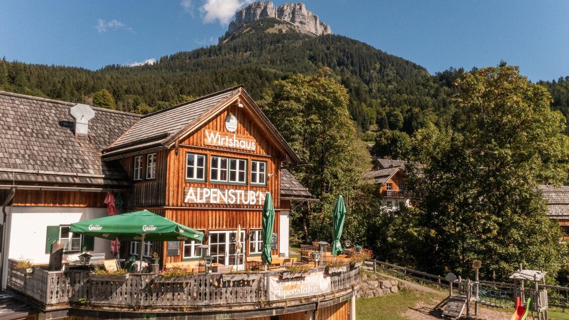 Ein gemütliches Wirtshaus mit einer Holzfassade und einer großen Terrasse. Im Hintergrund sind majestätische Berge und grüne Wälder zu sehen. | © Gerhard Wolkersdorfer
